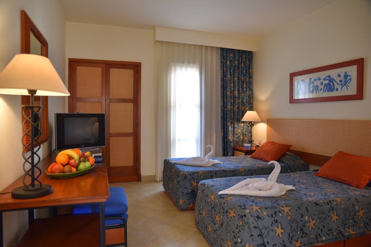 A hotel room with two beds, a desk with a fruit bowl, a television, and a framed picture on the wall.