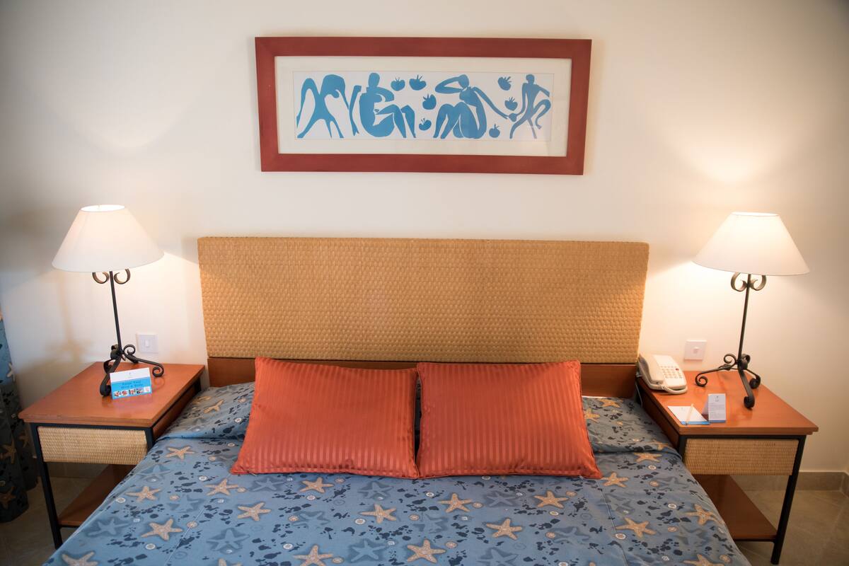 A hotel room with a bed featuring a blue star-patterned bedspread, two red pillows, a wooden headboard, and two bedside tables with lamps and a phone.