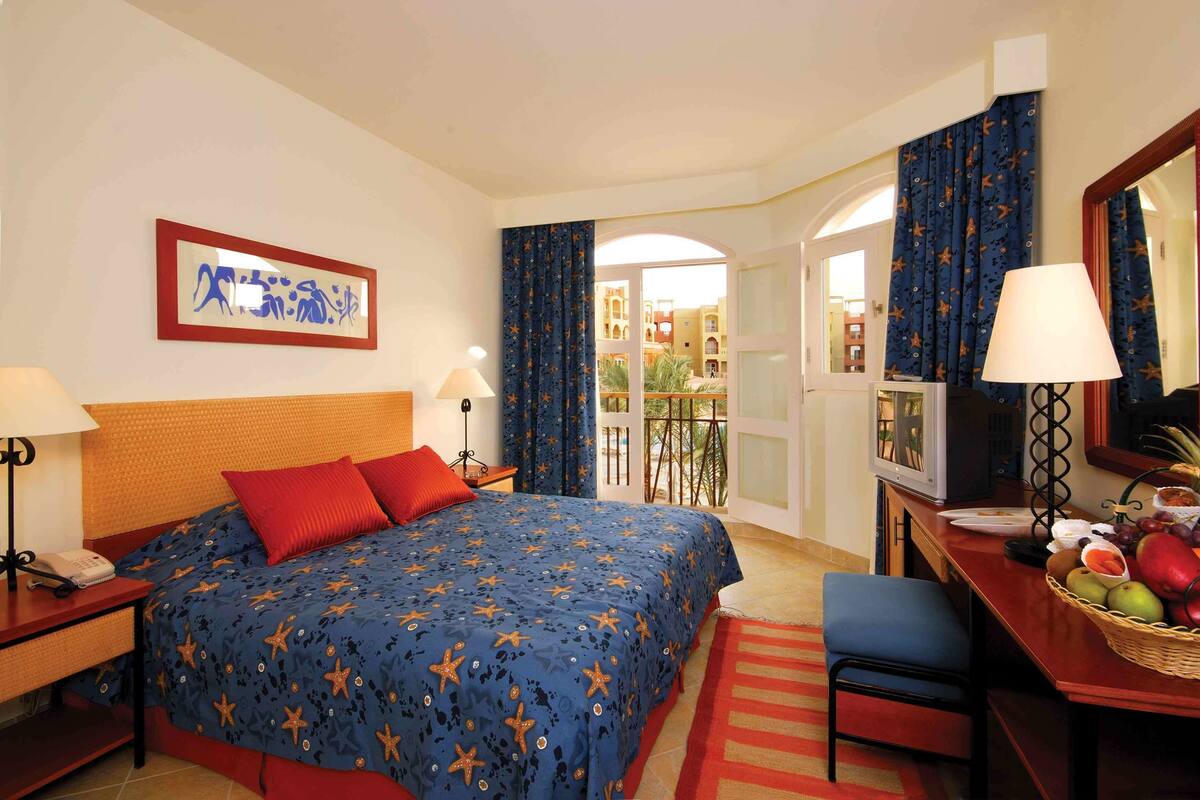 A hotel room with a bed, a desk with a lamp, a telephone, a fruit basket, and a view of the outside.