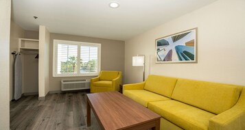 Una habitación con sofá amarillo, mesa de madera, ventana con persianas, radiador y un cuadro de rascacielos.