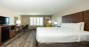 Habitación de hotel con cama, escritorio con televisor, silla y ventana con persianas.