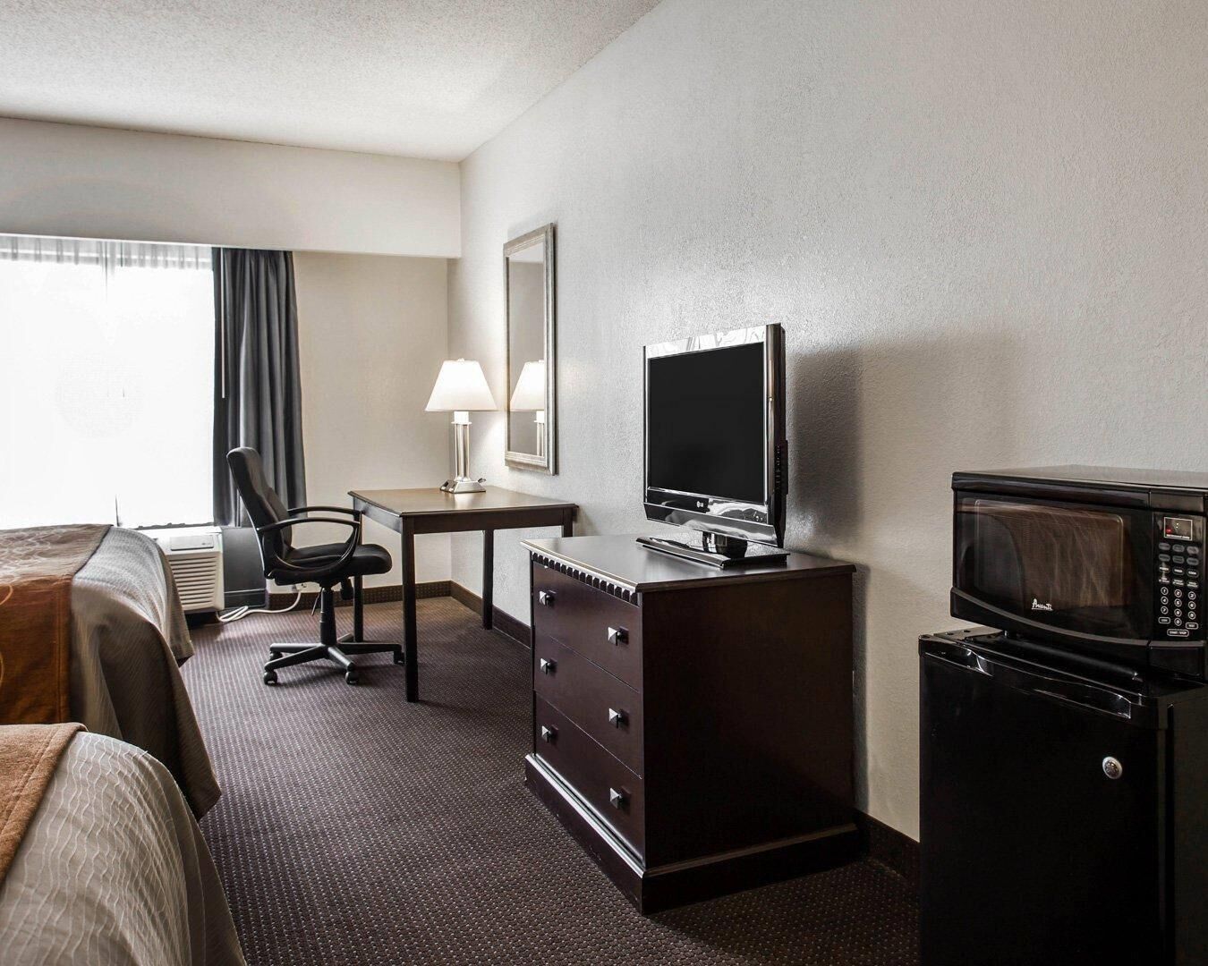 Une chambre d’hôtel comprenant un lit, un bureau avec un ordinateur, une chaise, une commode, un four à micro-ondes et une fenêtre avec des rideaux.