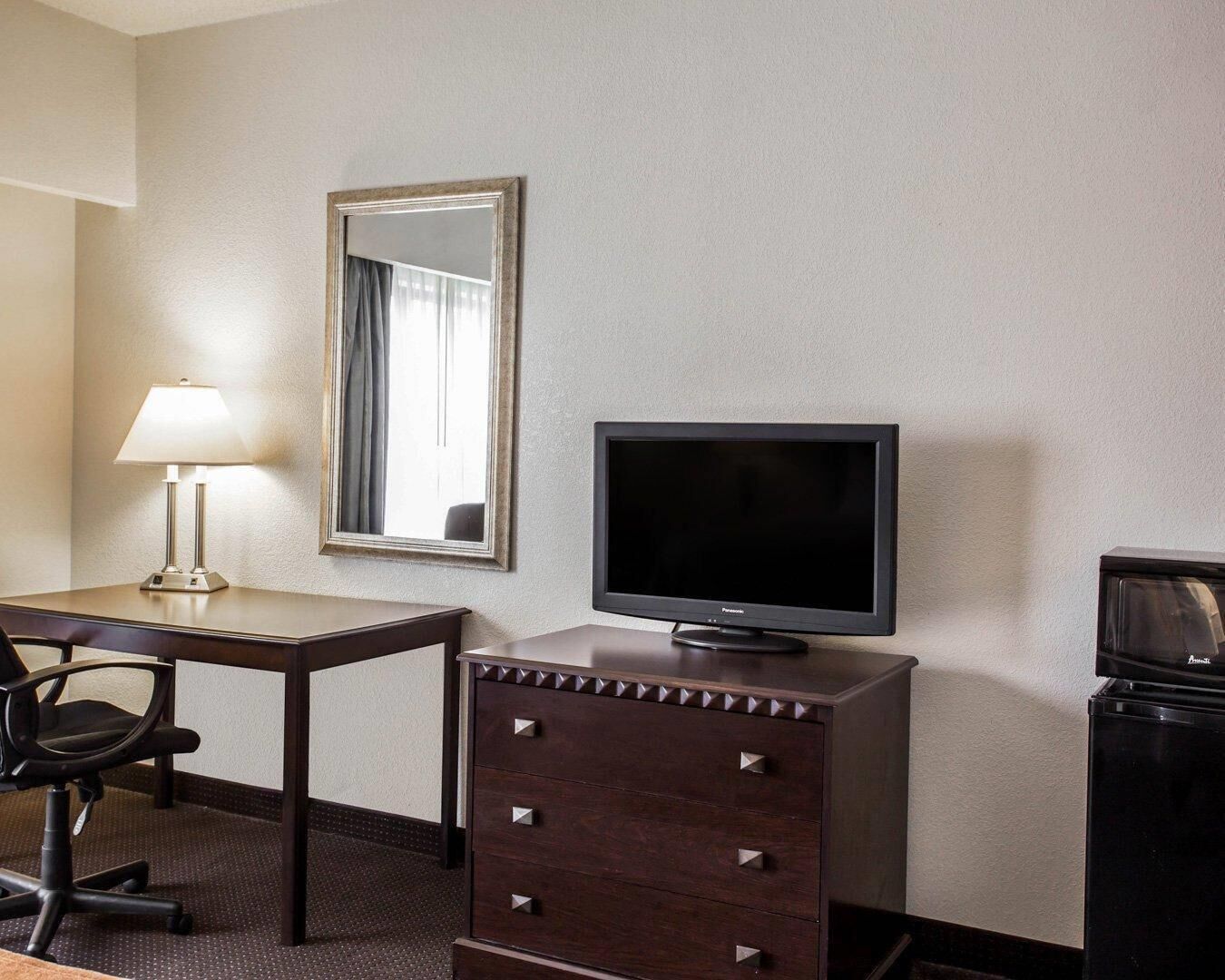 A hotel room with a desk, chair, TV, dresser, and a mirror.
