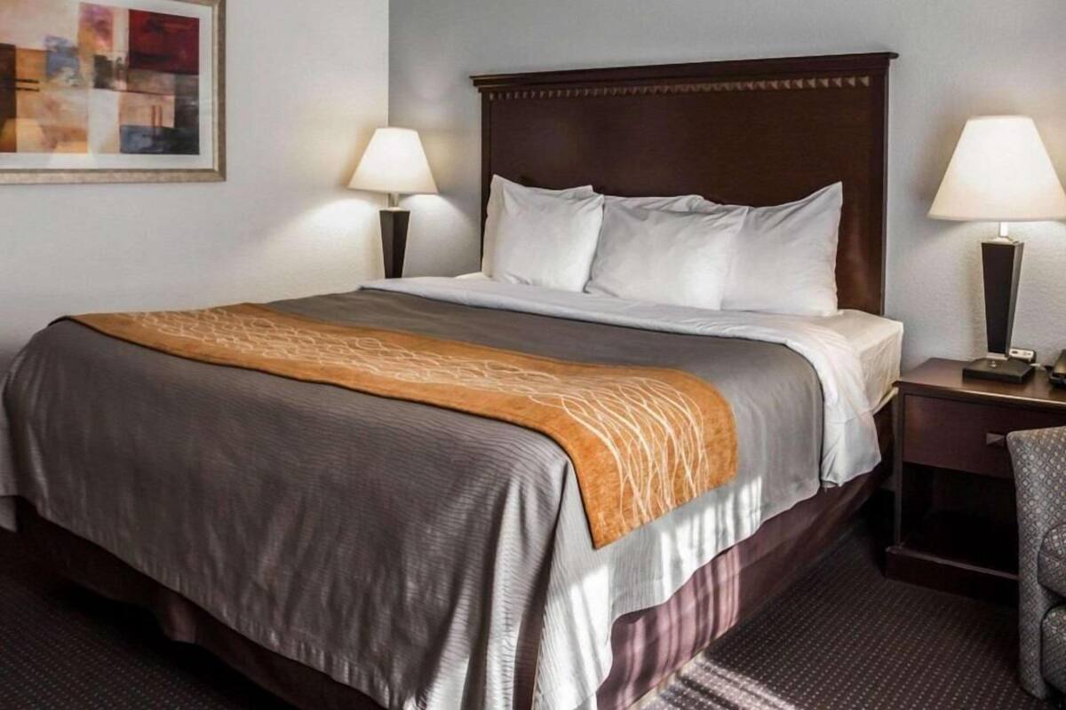 Una habitación de hotel con una cama grande, dos lámparas de noche, una mesita de noche, una silla y un cuadro en la pared.