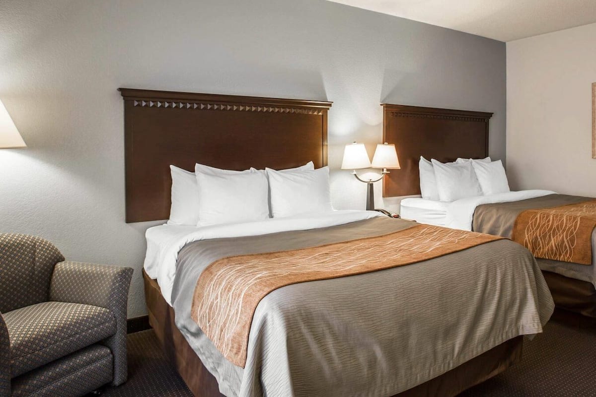 Habitación de hotel con dos camas, una mesita de noche con lámparas, una silla y un papel tapiz estampado.