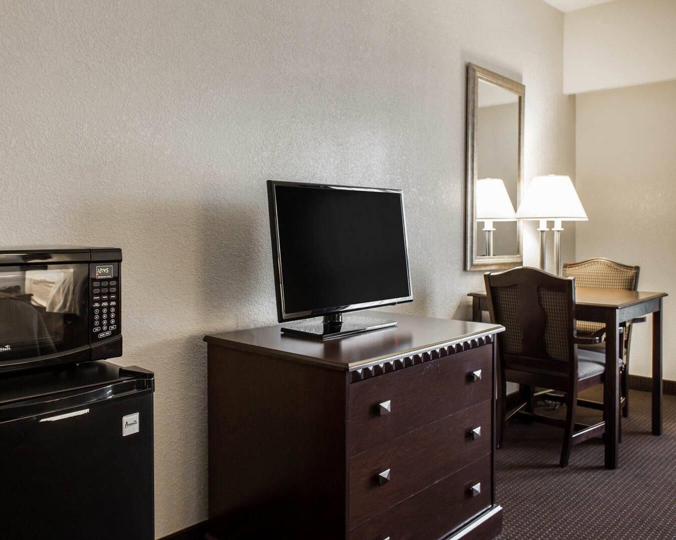 A hotel room with a flat-screen TV on a dresser, a microwave, a desk with a chair, and a lamp.