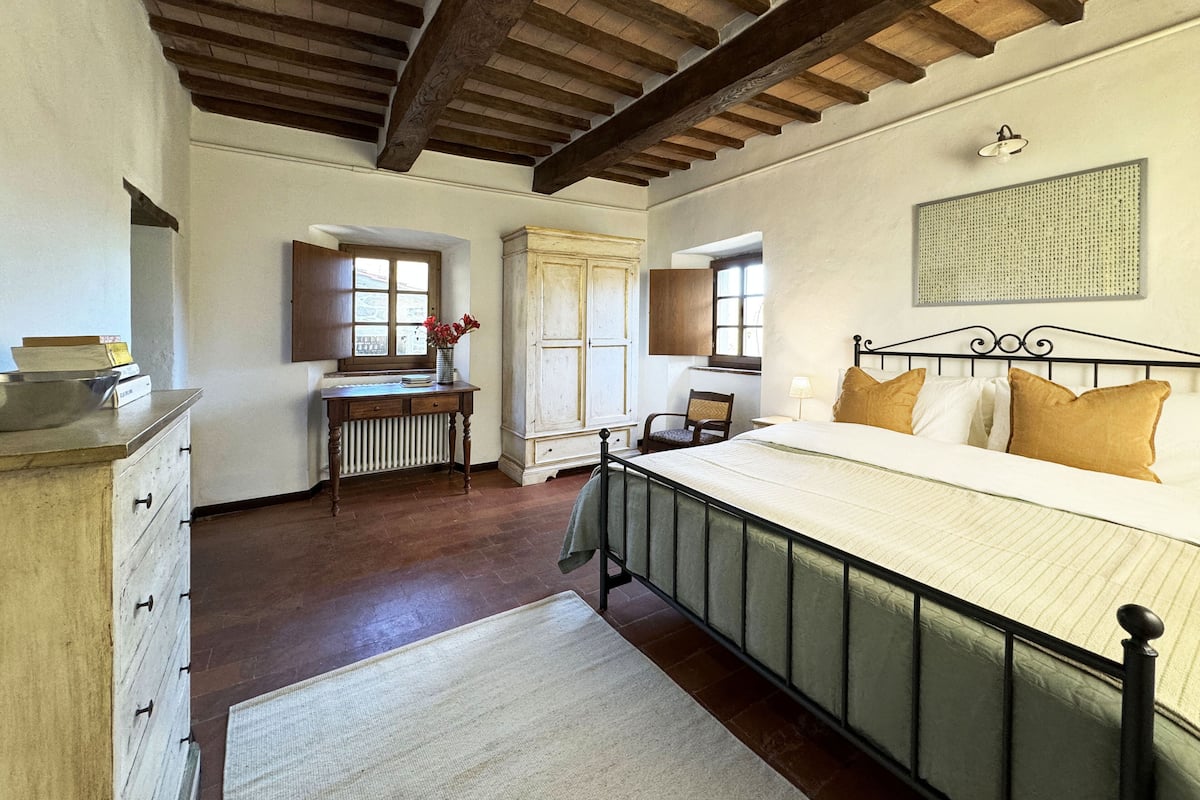 A bedroom with a wooden ceiling, a bed with a metal frame, a radiator, a window, and a chair.