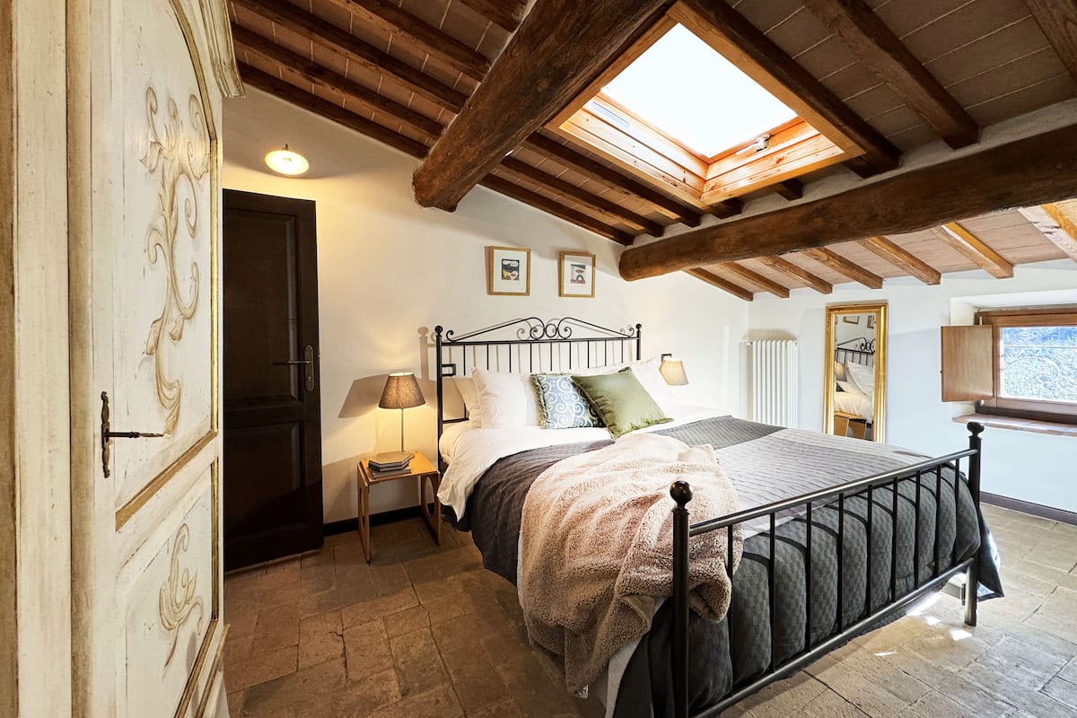 A bedroom with a sloped ceiling, a skylight, a bed with a metal frame, a nightstand with a lamp, and a wooden door with decorative moldings.