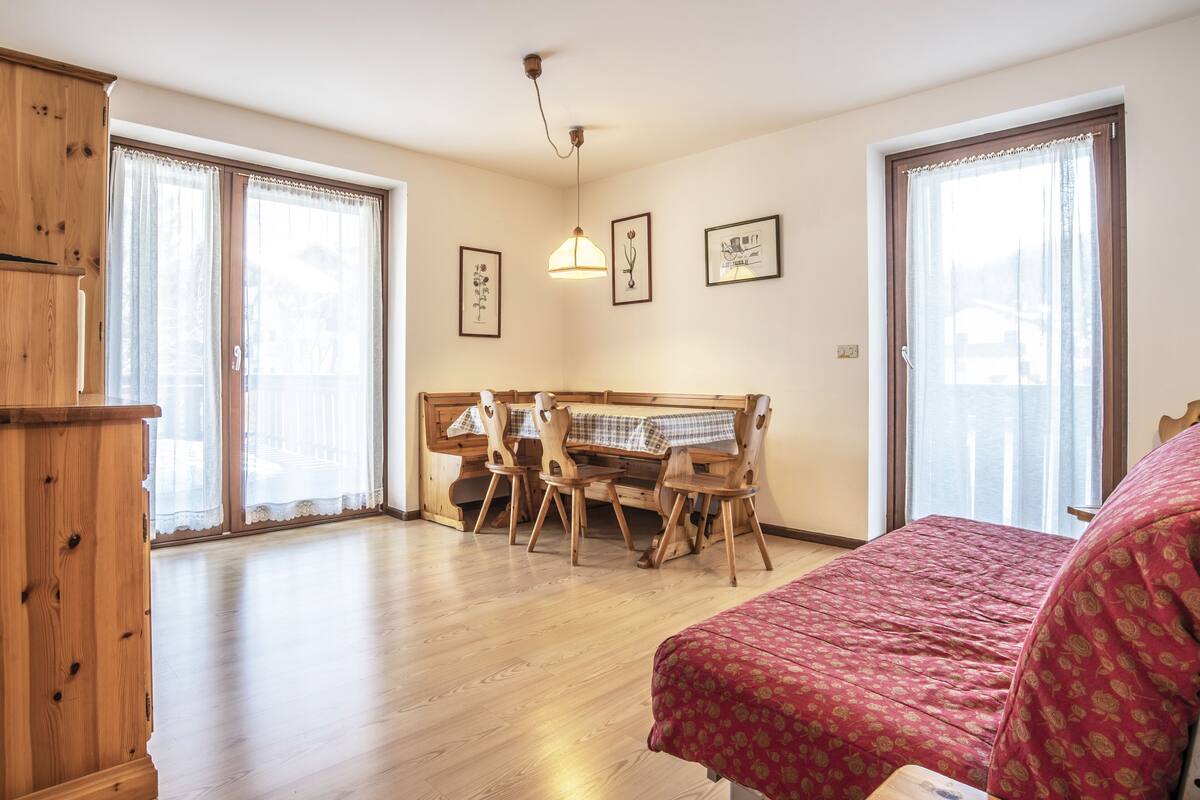 A spacious room with a wooden floor, a bed with a patterned bedspread, a wooden bench table with chairs, and large windows with sheer curtains.