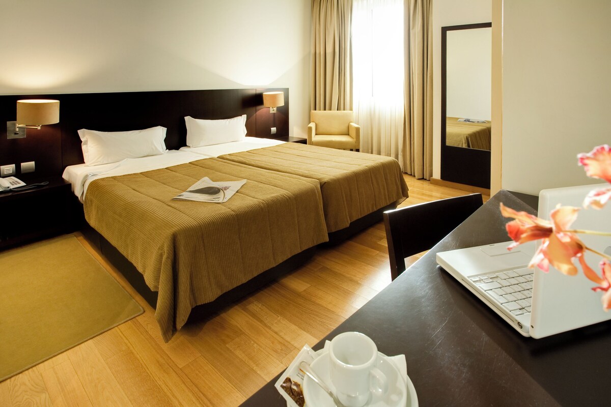 Una habitación de hotel con una cama, una silla, un escritorio con una computadora portátil y una taza en el escritorio.