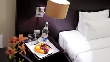 Una cama bien hecha con sábanas blancas, una mesita de noche con una bandeja de fruta y una botella de agua, y una lámpara en la pared.