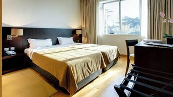 Quarto de hotel com uma cama grande, uma escrivaninha e uma cadeira. Há uma janela com cortinas, uma mesa de cabeceira com uma luminária e um telefone na mesa de cabeceira.
