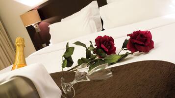 A neatly made bed with white linens, a bottle of champagne, two wine glasses, and three red roses on the bed.