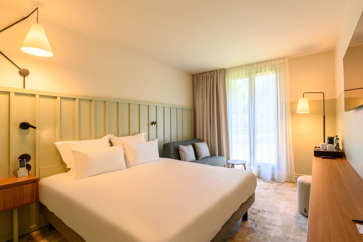 A hotel room with a large bed, a sofa, a desk, a TV, and a window with curtains.