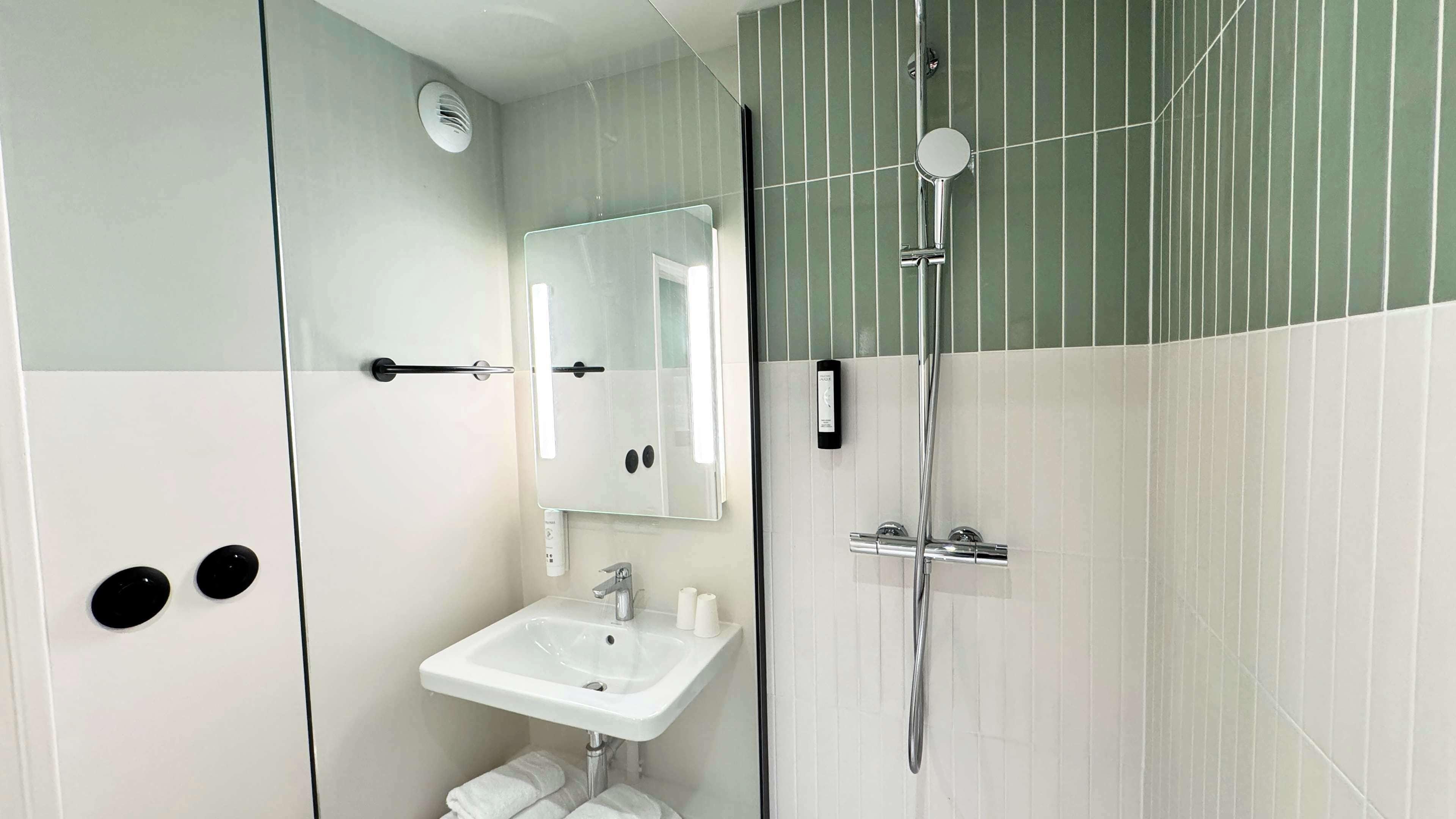 Un baño moderno con un lavamanos blanco, un espejo, un cabezal de ducha manual y un perchero para toallas.