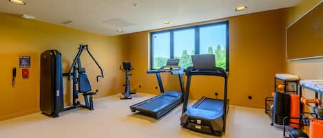 Ein Fitnessraum mit Trainingsgeräten, einem Laufband und einem Fenster mit Blick auf Bäume.