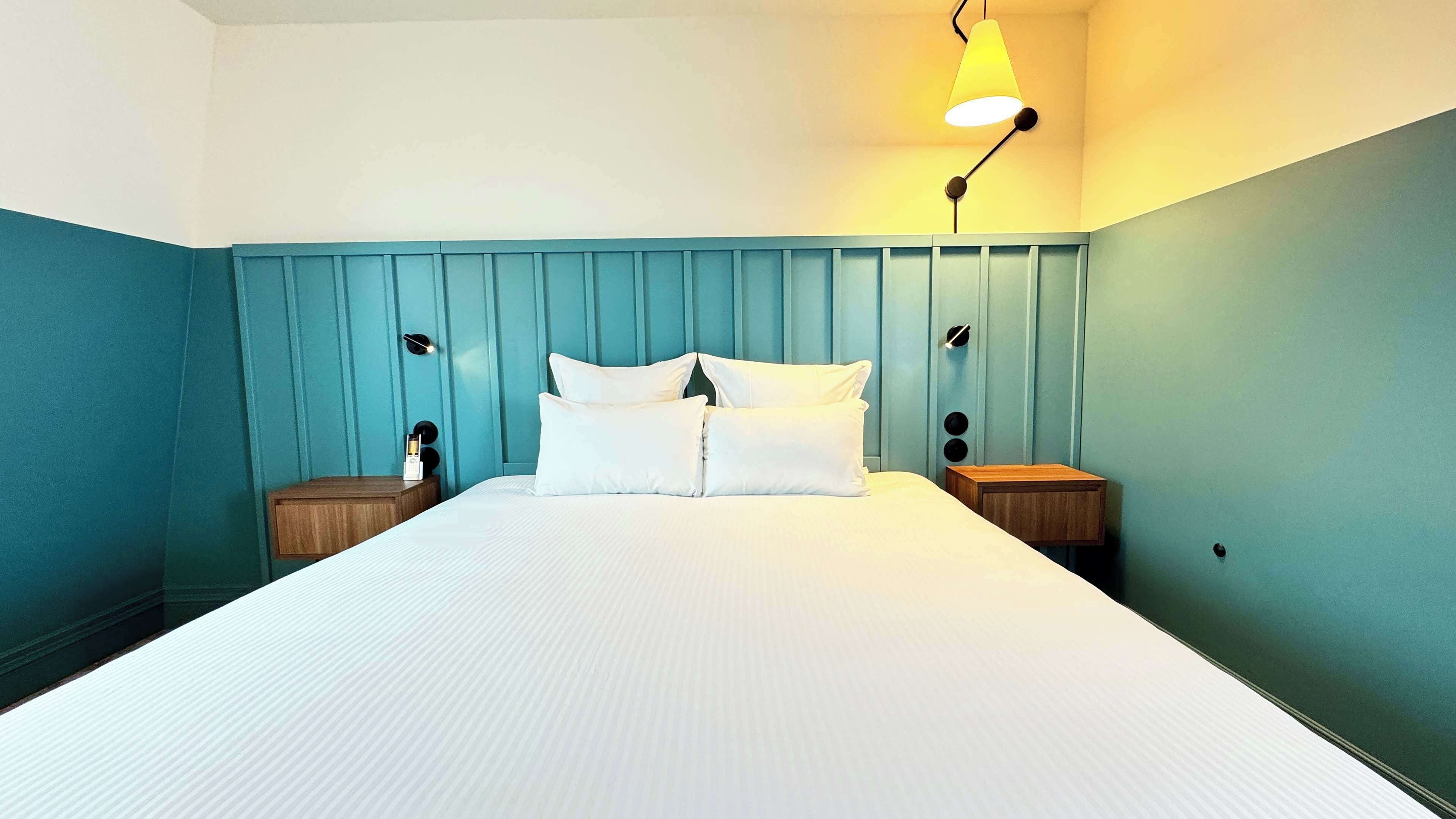 Una cama bien hecha con sábanas blancas en una habitación con paredes verde azulado y un cabecero de madera. Hay dos mesitas de noche con lámparas y un pequeño adorno sobre una de ellas.