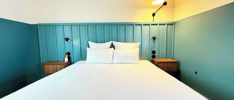 Una cama bien hecha con sábanas blancas en una habitación con paredes verde azulado y un cabecero de madera. Hay dos mesitas de noche con lámparas y un pequeño adorno sobre una de ellas.