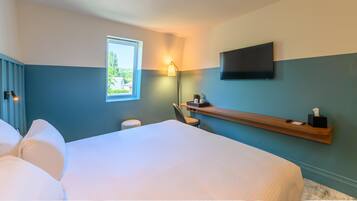 A hotel room with a large bed, a desk with a chair, a TV mounted on the wall, and a window with a view of greenery.