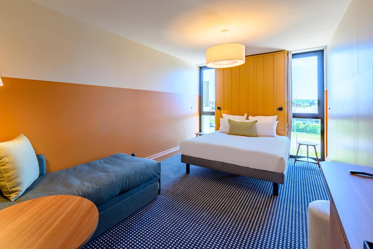 A modern hotel room with a bed, a sofa, a small table, and a large window offering a view.