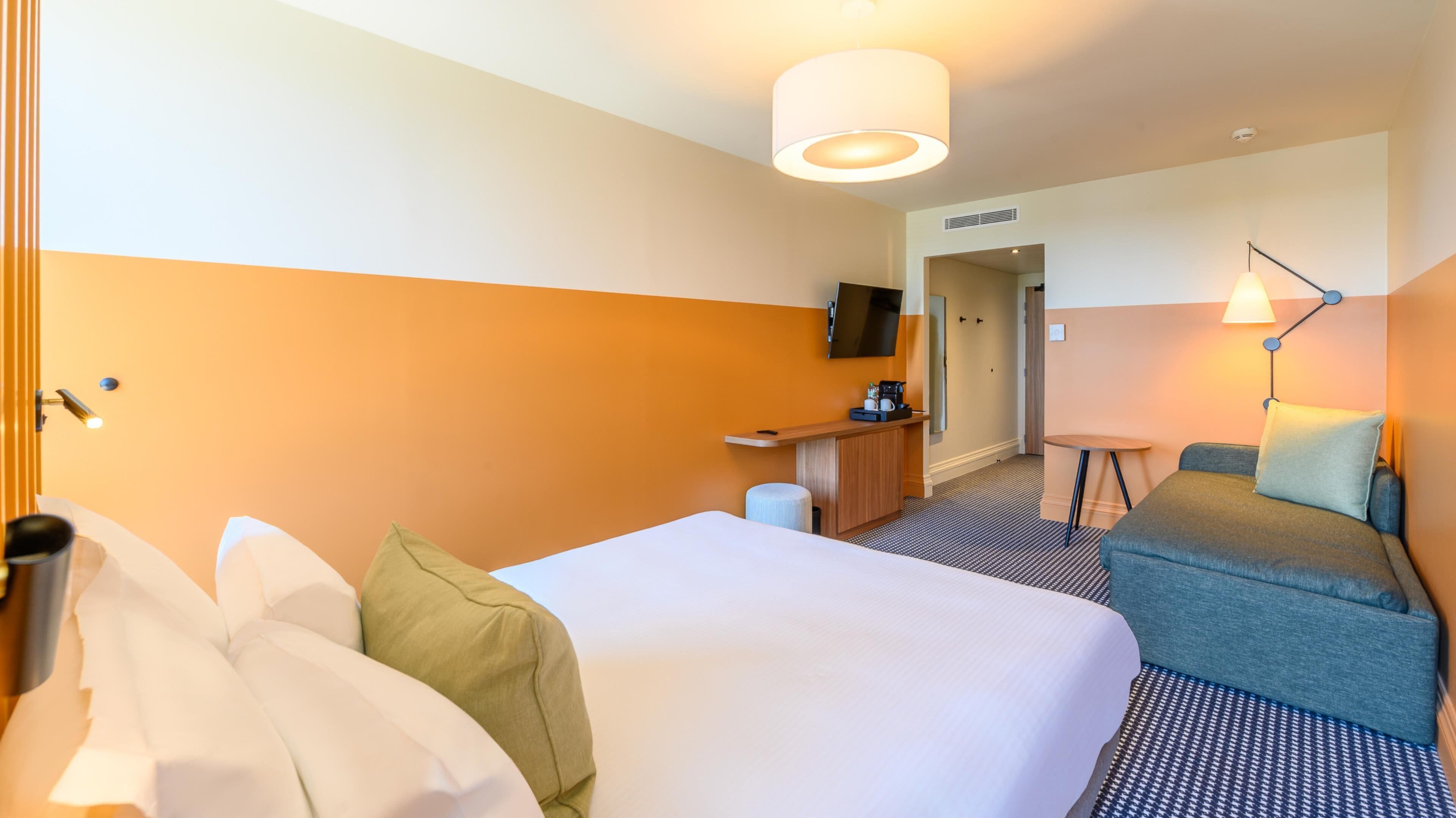 Una habitación de hotel con una cama grande, un escritorio con una silla, una mesita de noche con una lámpara y un televisor en la pared.