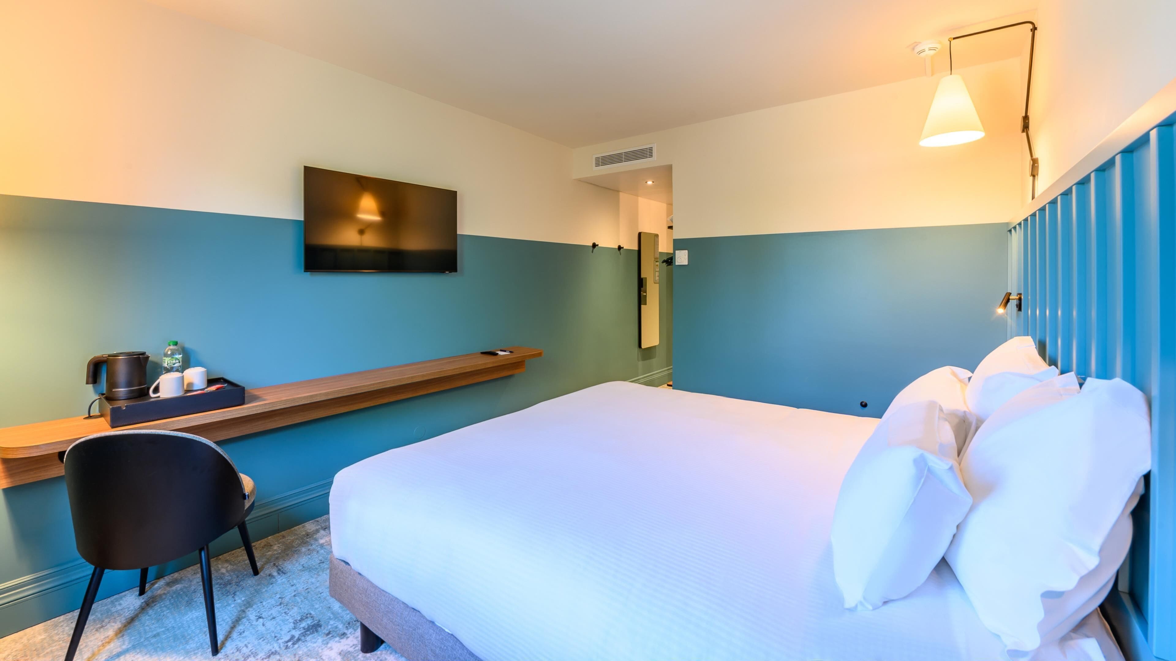 A modern hotel room with a bed, a desk with a kettle and mugs, a chair, and a TV mounted on the wall.
