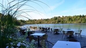 Un área de comedor al aire libre con mesas y sillas frente a un lago, rodeada de vegetación.