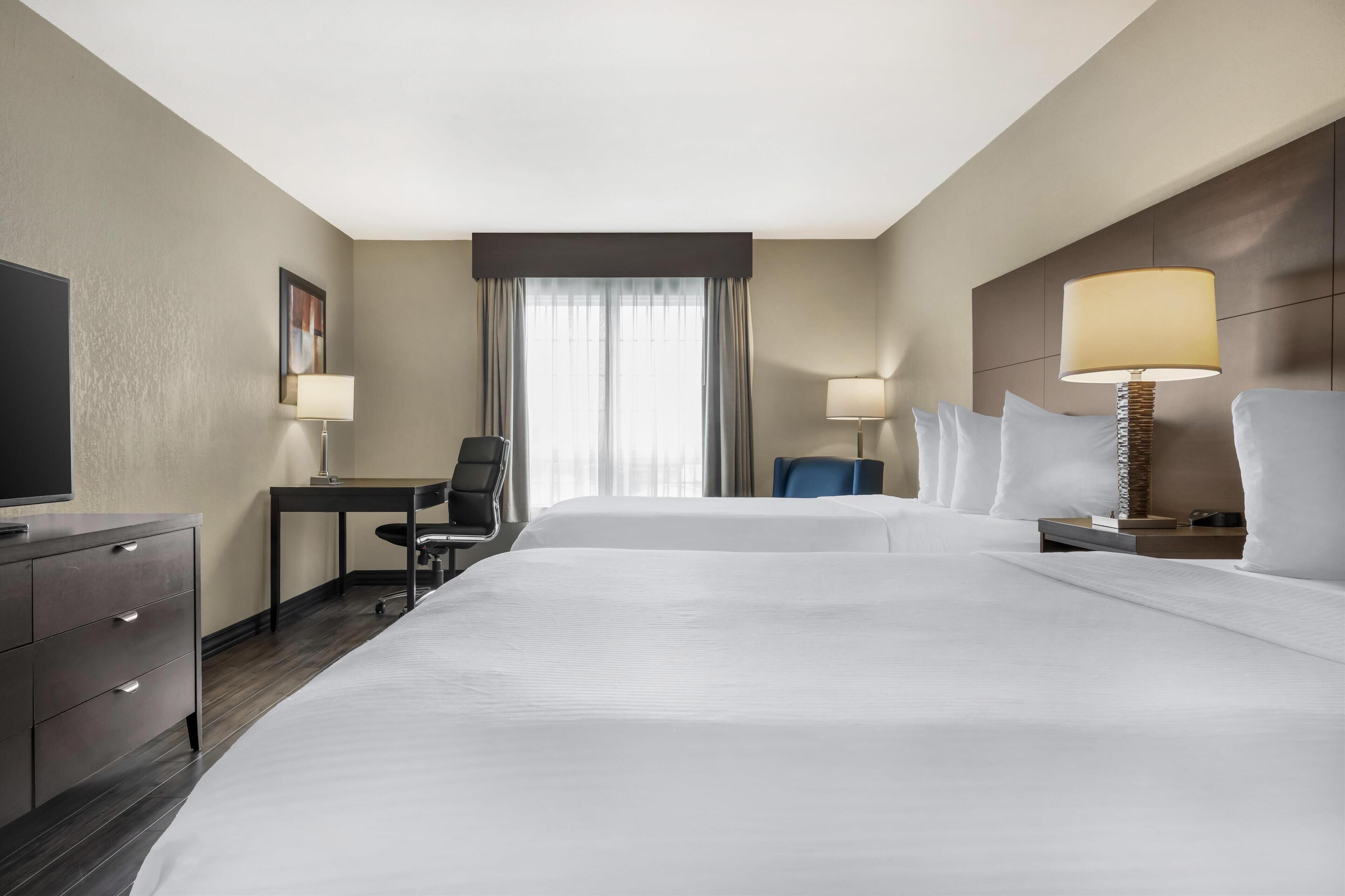 A hotel room with two beds, a desk, a chair, a TV, and two lamps.