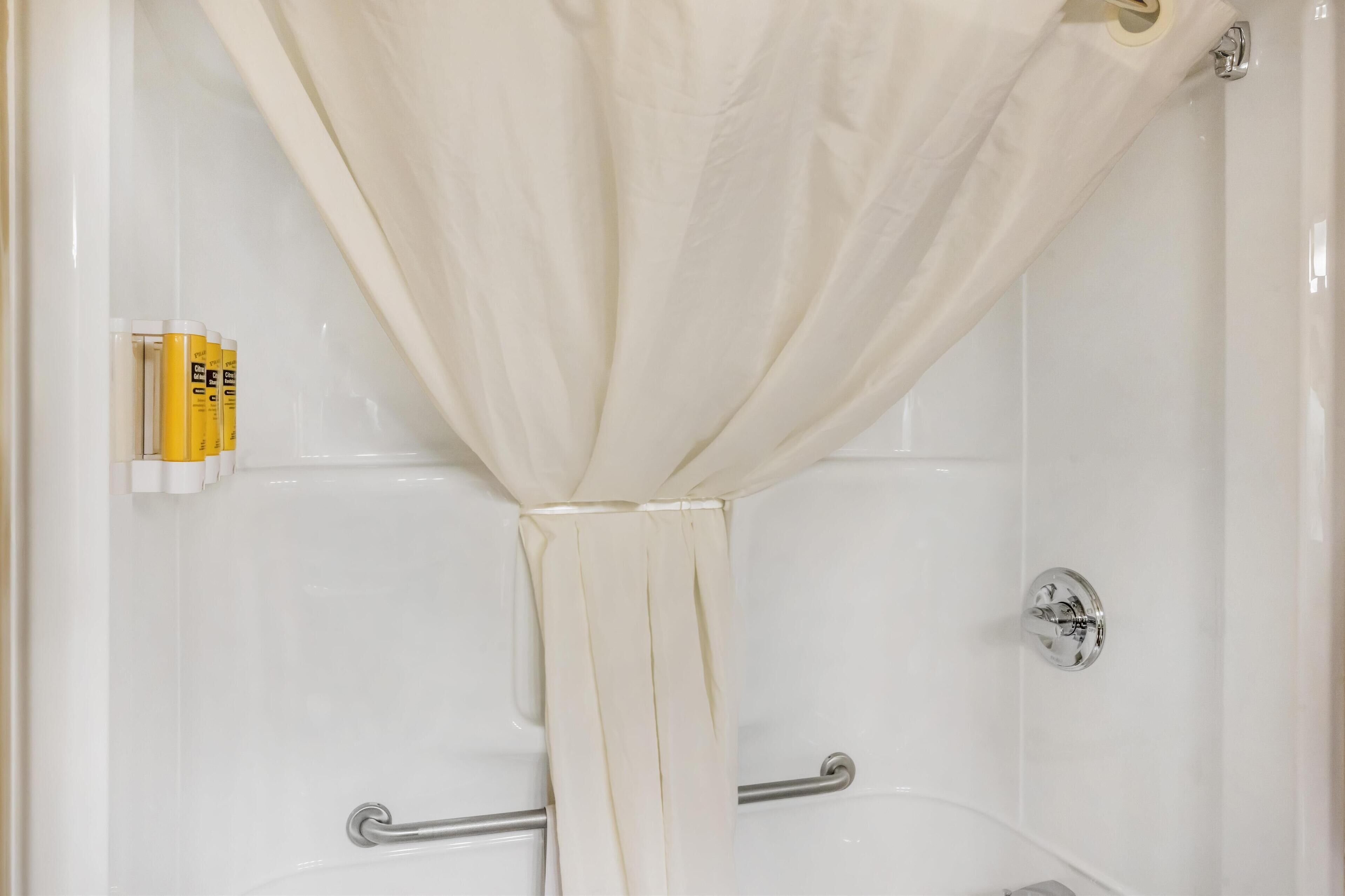 Une salle de bain avec un rideau de douche attaché en haut, une barre d’appui et un ensemble de bouteilles jaunes et blanches fixées au mur.