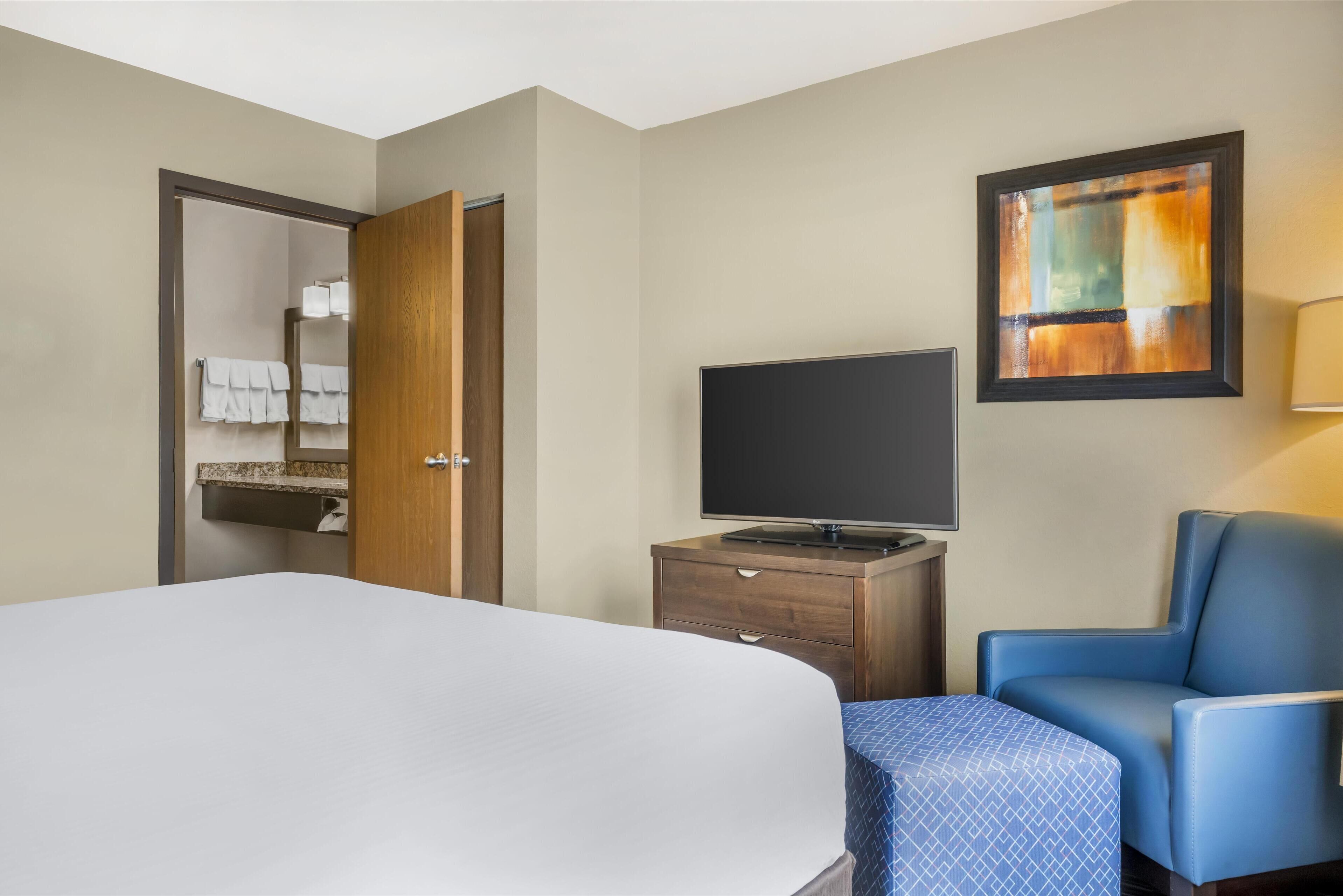 Een hotelkamer met een bed, een televisie op een houten dressoir, een blauwe fauteuil en een schilderij aan de muur.