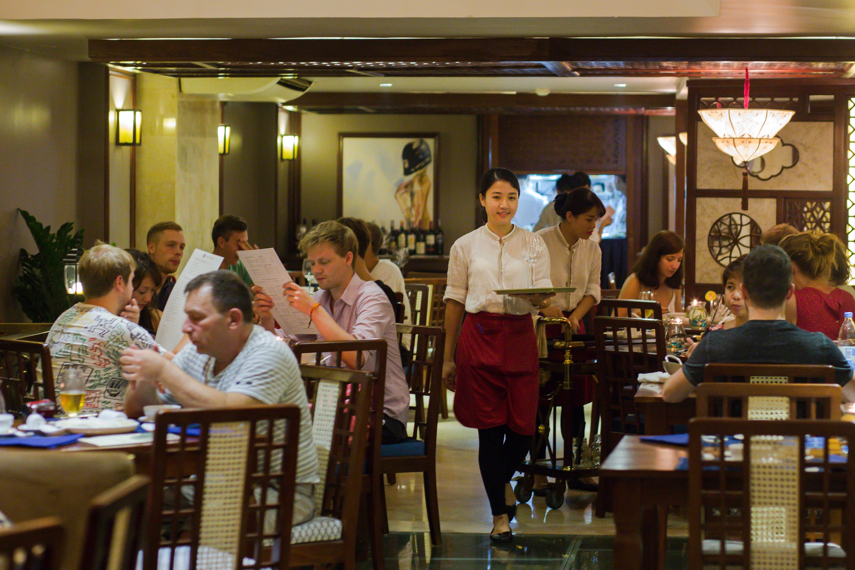 A restaurant with wooden chairs and tables, a waiter in uniform, and patrons dining.