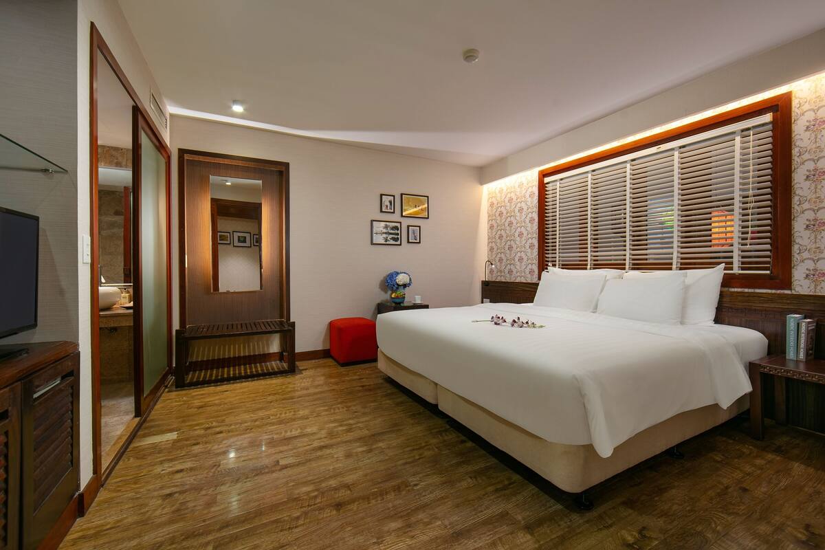 Une chambre d’hôtel avec un grand lit, un plancher en bois, une télévision, un pouf rouge et une fenêtre avec des stores.