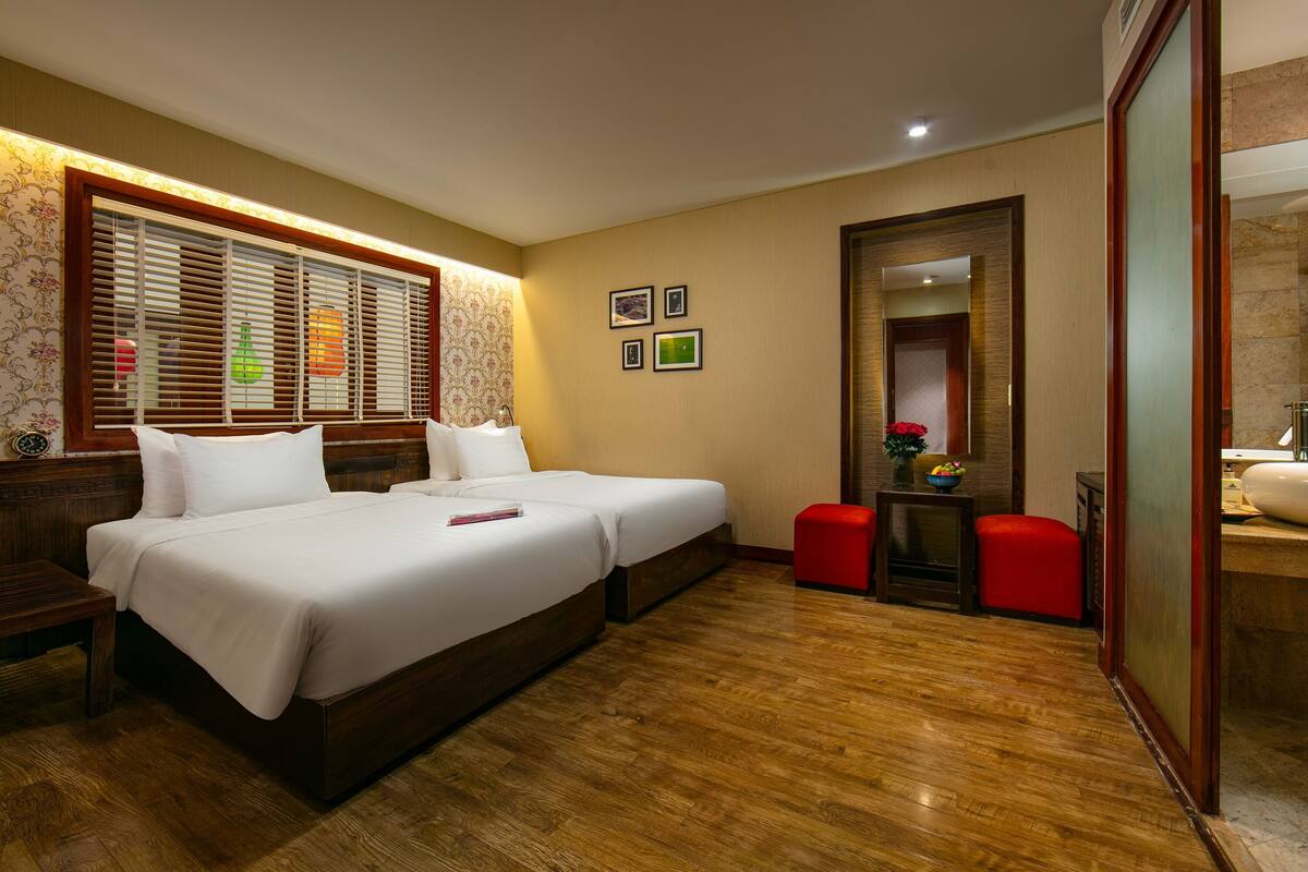 Une chambre d’hôtel avec un grand lit, deux tables de chevet, un petit tabouret rouge, un plancher en bois et une salle de bain visible à travers une porte en verre.