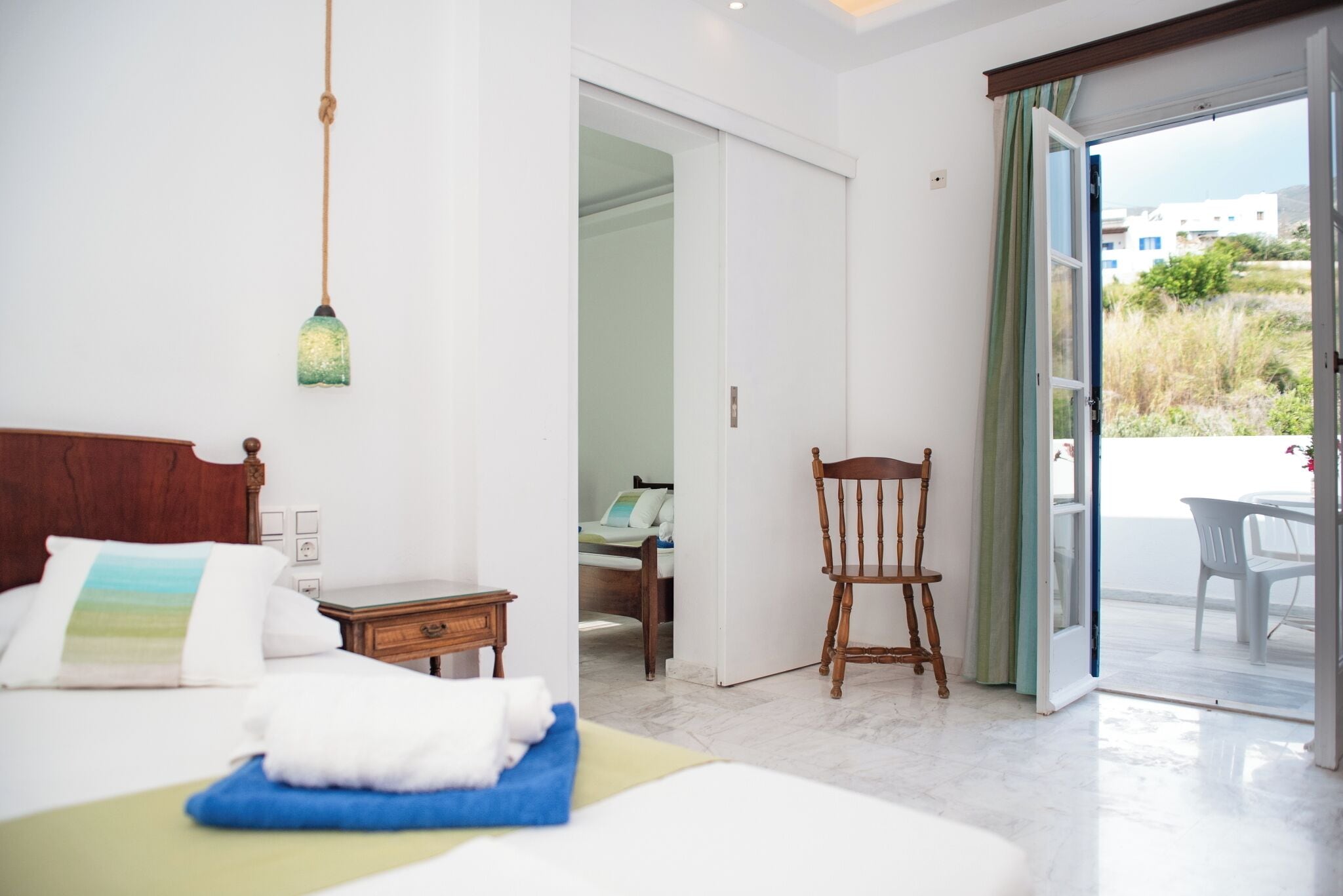 Un dormitorio con cama, mesita de noche, silla y vistas a un balcón desde una puerta abierta.