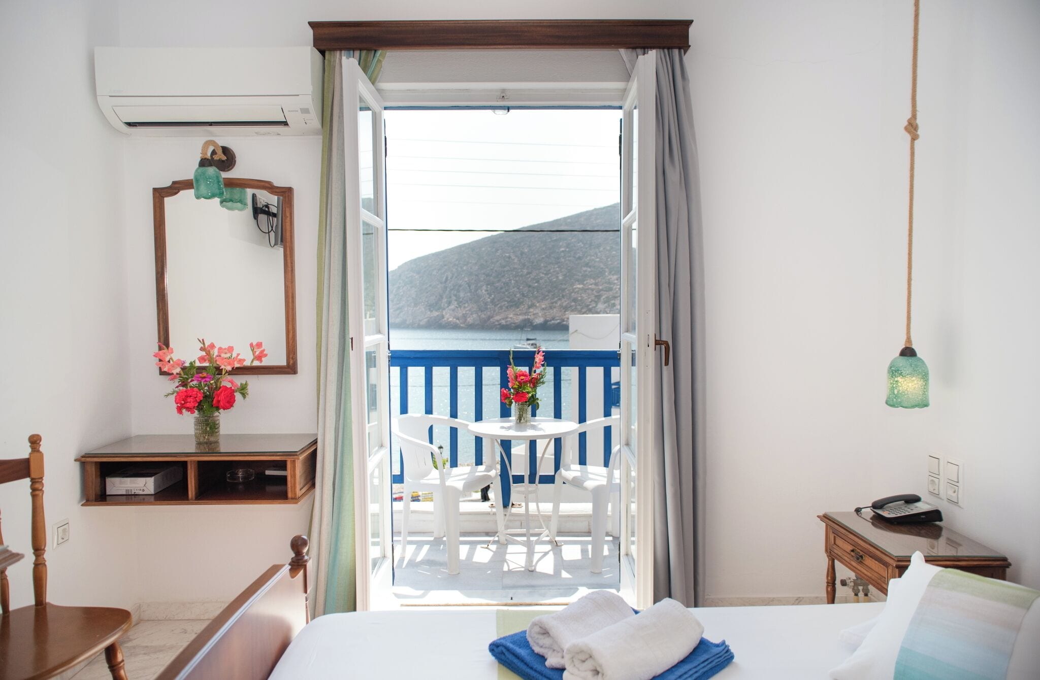 Habitación de hotel con cama, escritorio, silla, una pequeña mesa con un florero y vista al mar desde el balcón.