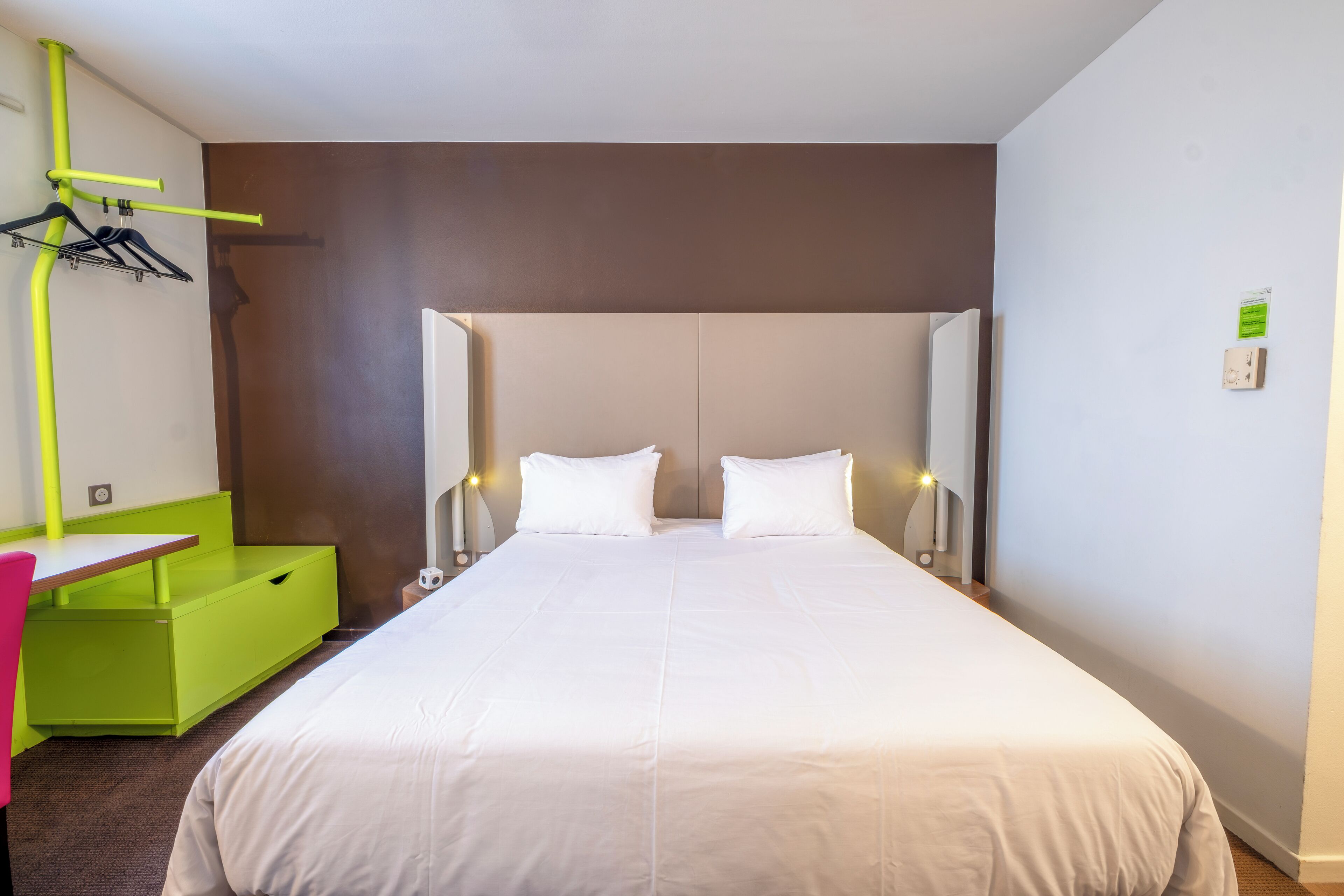 A hotel room with a large bed, a desk with a chair, and a green bench.