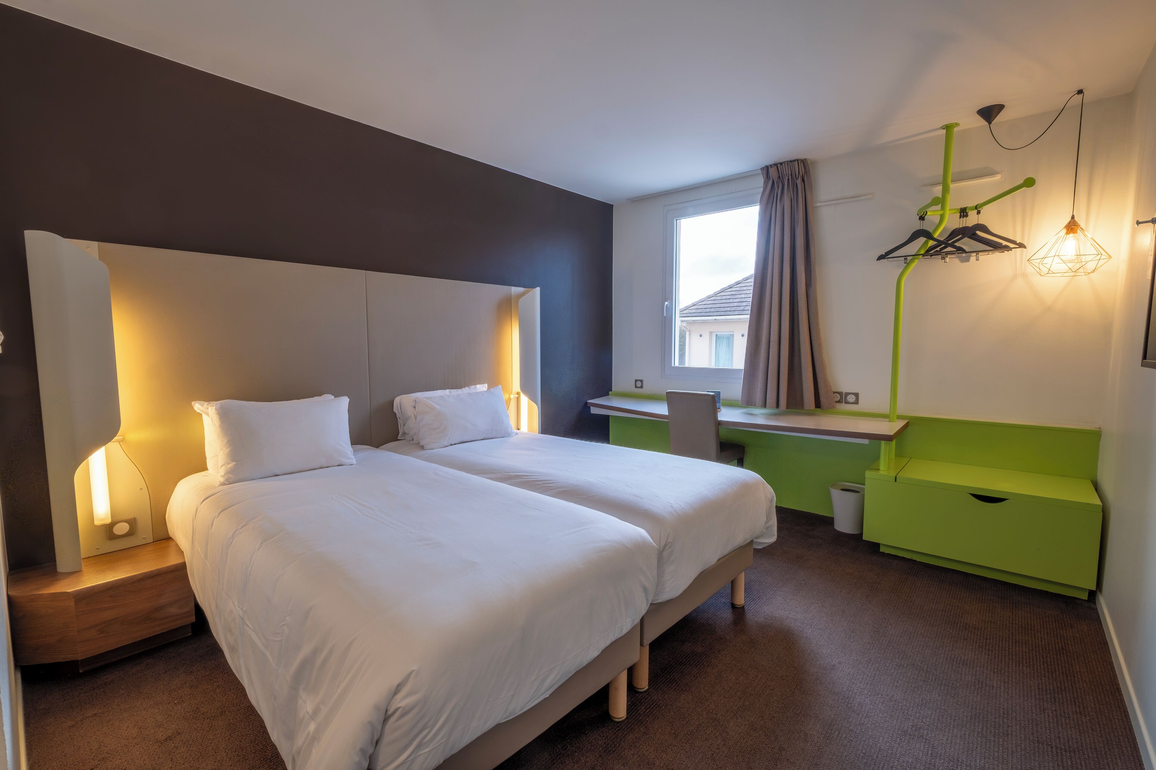 A hotel room with two beds, a desk, a chair, a green dresser, and a window with curtains.