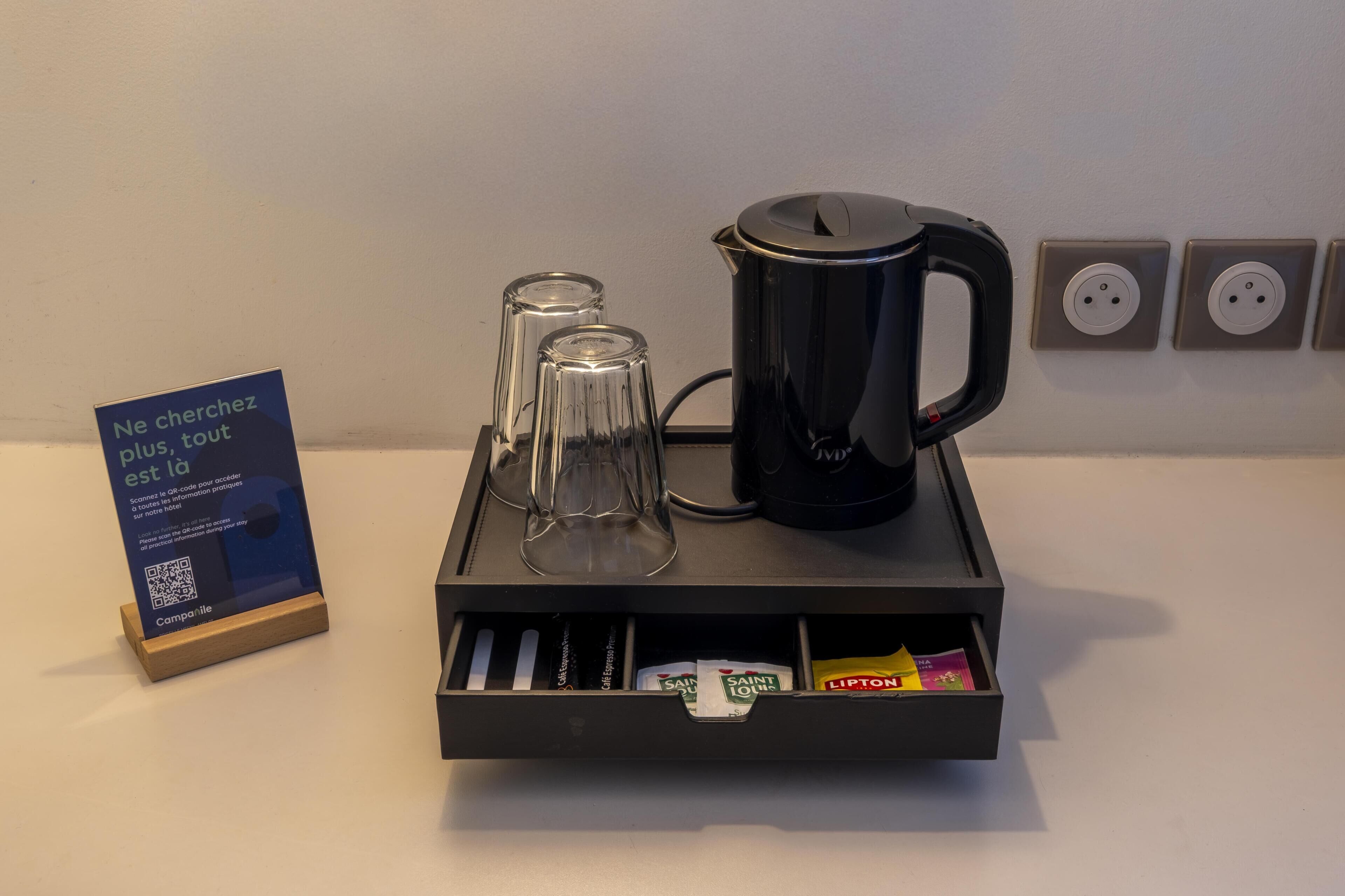 Una estación de café con una cafetera negra, dos vasos transparentes y un cajón con sobrecitos de café.