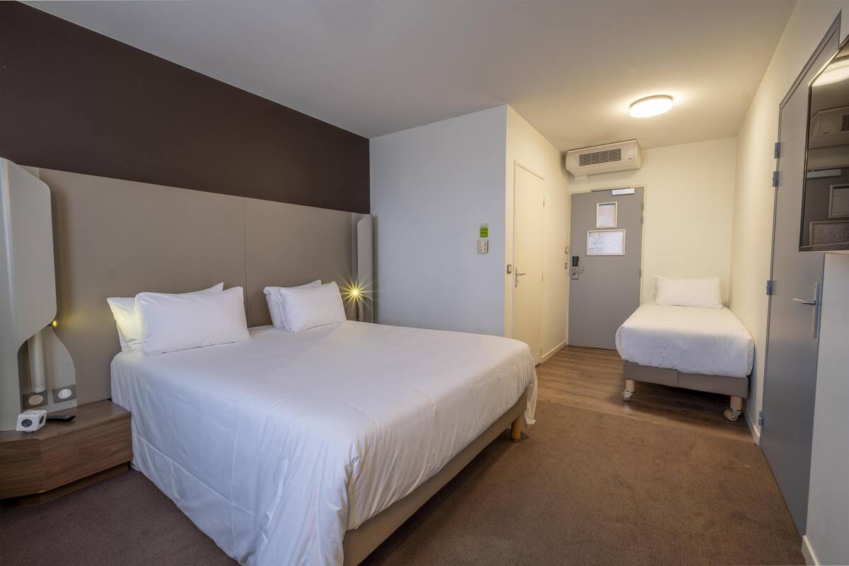 Quarto de hotel com uma cama grande, uma cama menor, uma mesinha de cabeceira e um ar condicionado fixado na parede.