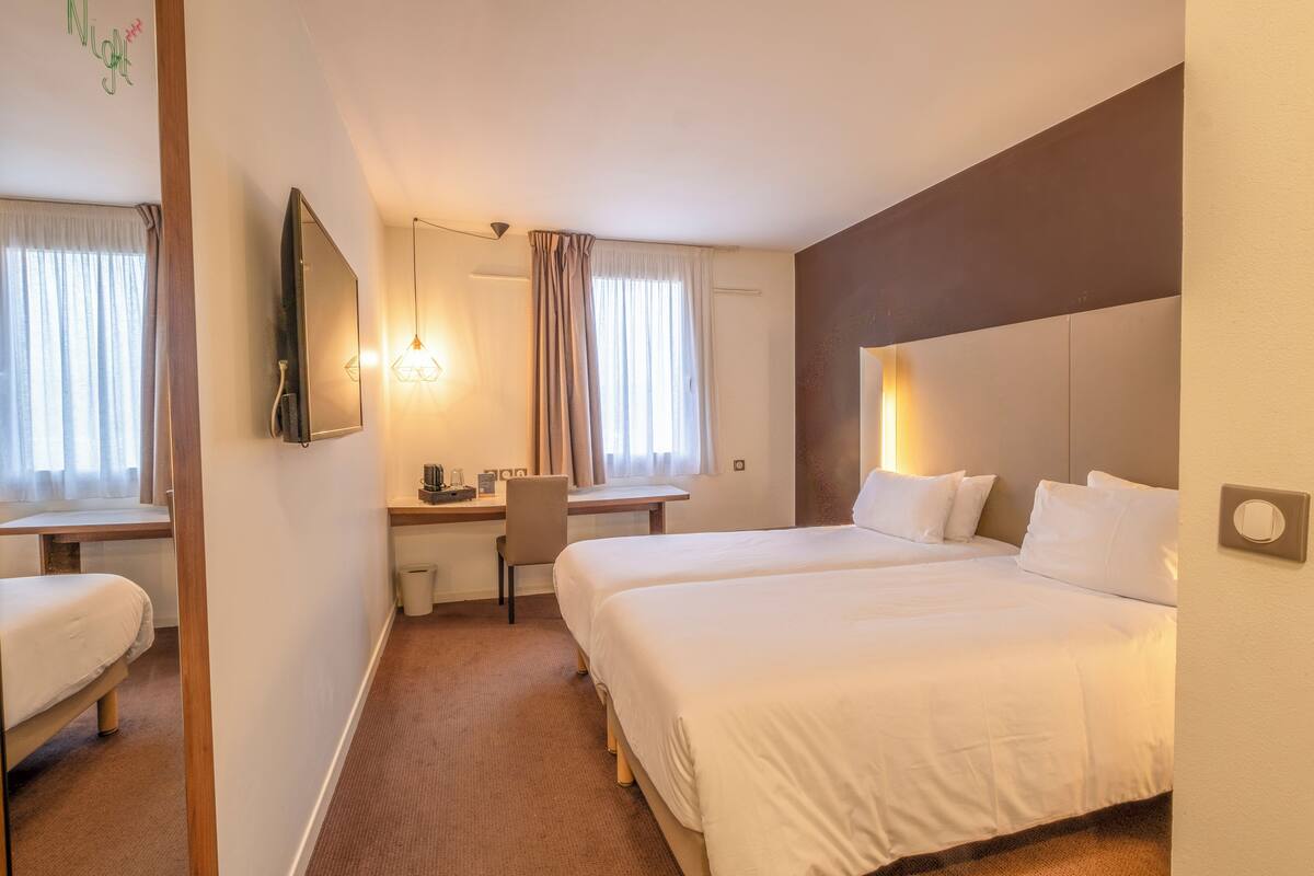 Quarto de hotel com duas camas, uma escrivaninha, uma cadeira, televisão e janela com cortinas.