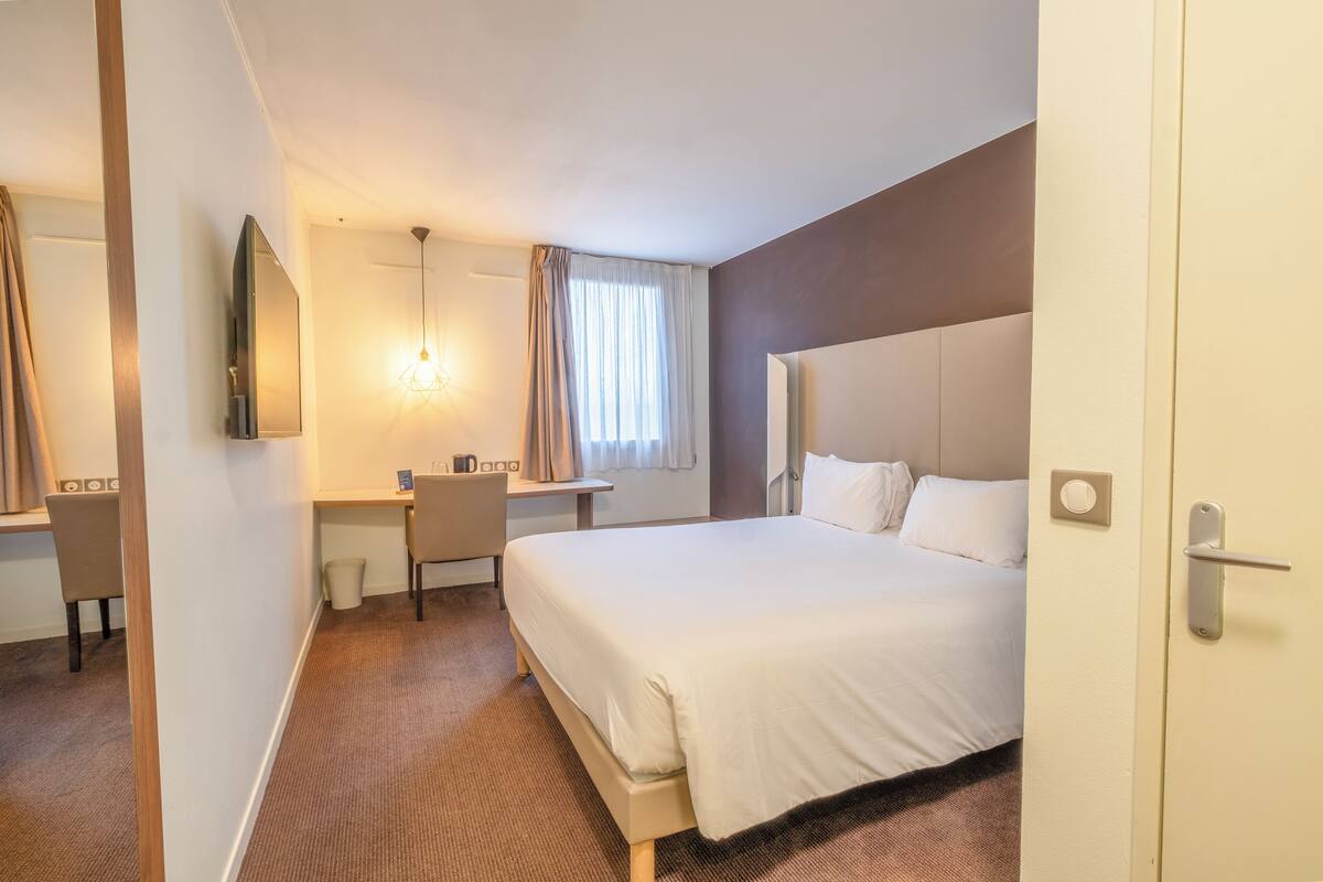 Quarto de hotel com cama, escrivaninha, cadeira, televisão e janela com cortinas.