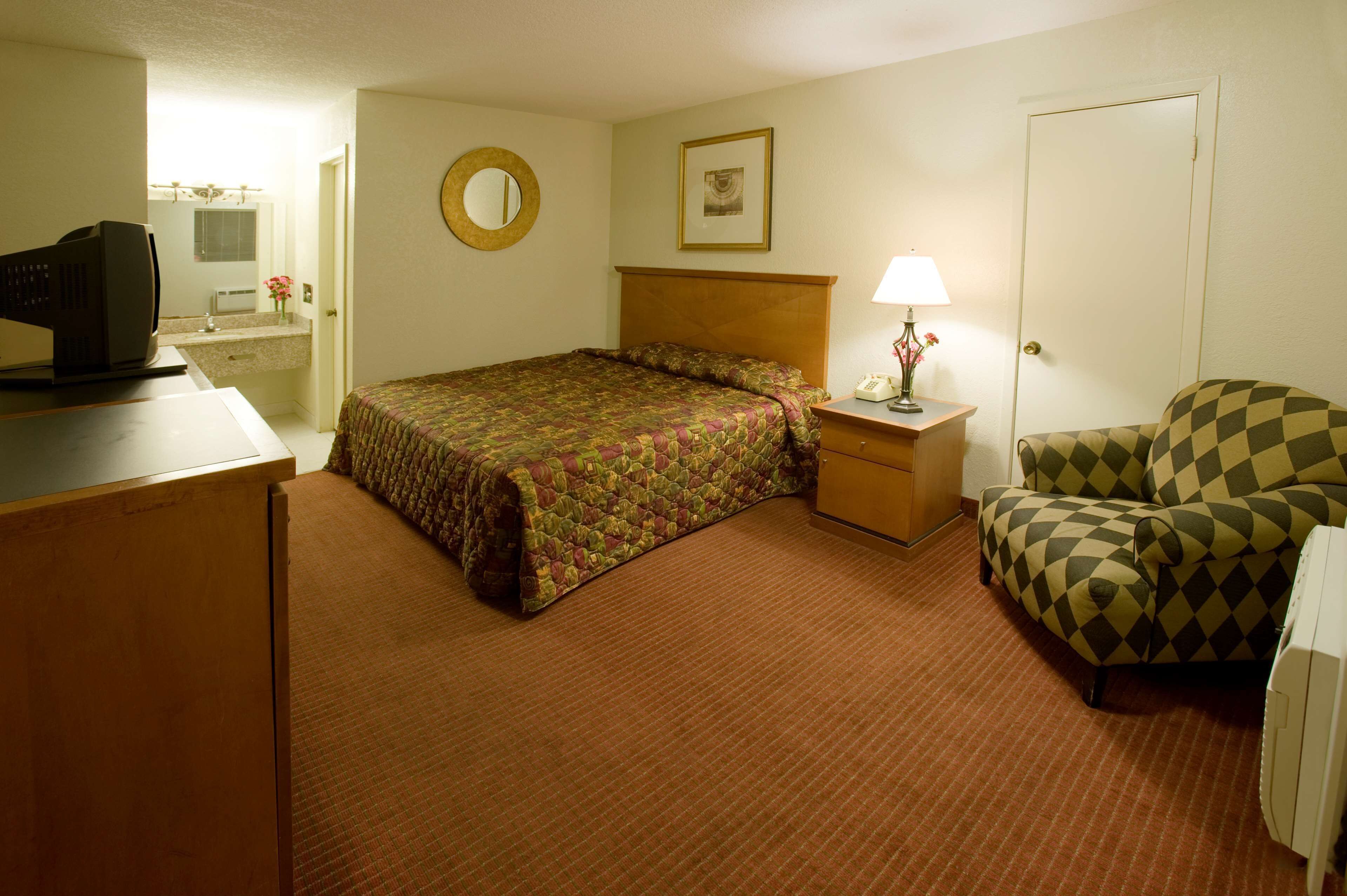 A hotel room with a bed, a desk with a computer, a chair, a nightstand with a lamp, and a door.