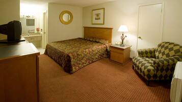 A hotel room with a bed, a desk with a computer, a chair, a nightstand with a lamp, and a door.