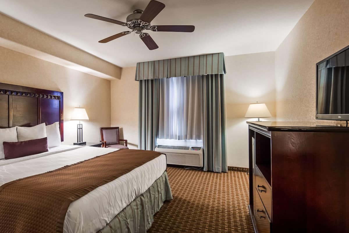 A hotel room with a large bed, a ceiling fan, a TV, a desk with a lamp, and a window with curtains.
