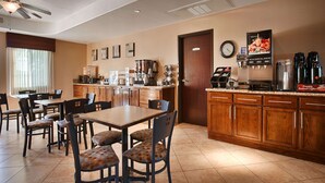 A hotel dining area with a kitchen in the background, featuring a coffee machine, a clock, and a window with curtains. - Best Western Plus Main Street Inn (Brawley)