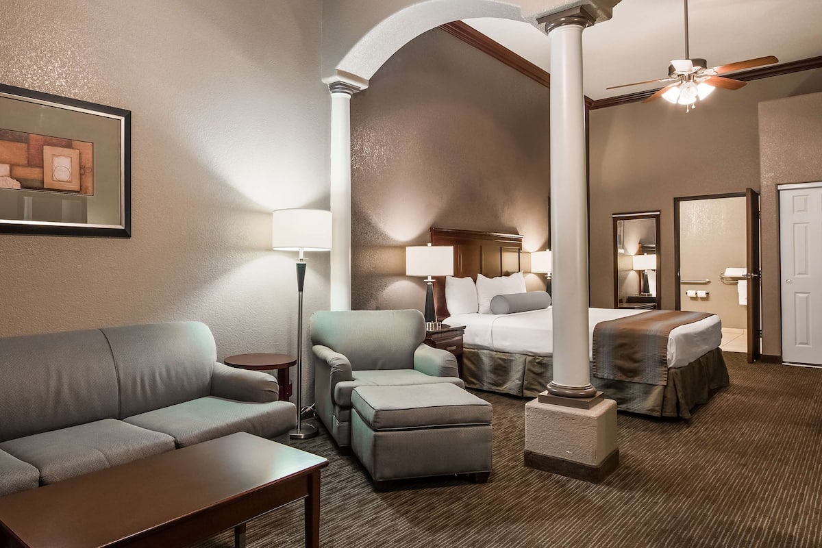 A hotel room with a sofa, armchair, ottoman, a bed, a nightstand, a lamp, a ceiling fan, and a framed picture on the wall.