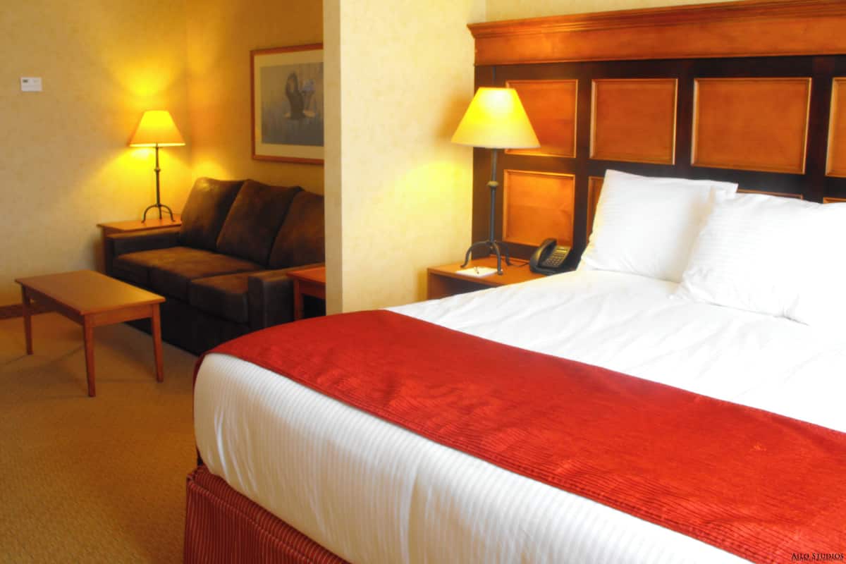 A hotel room with a large bed, a wooden headboard, a nightstand with a lamp, a small table, a sofa, and a framed picture on the wall.