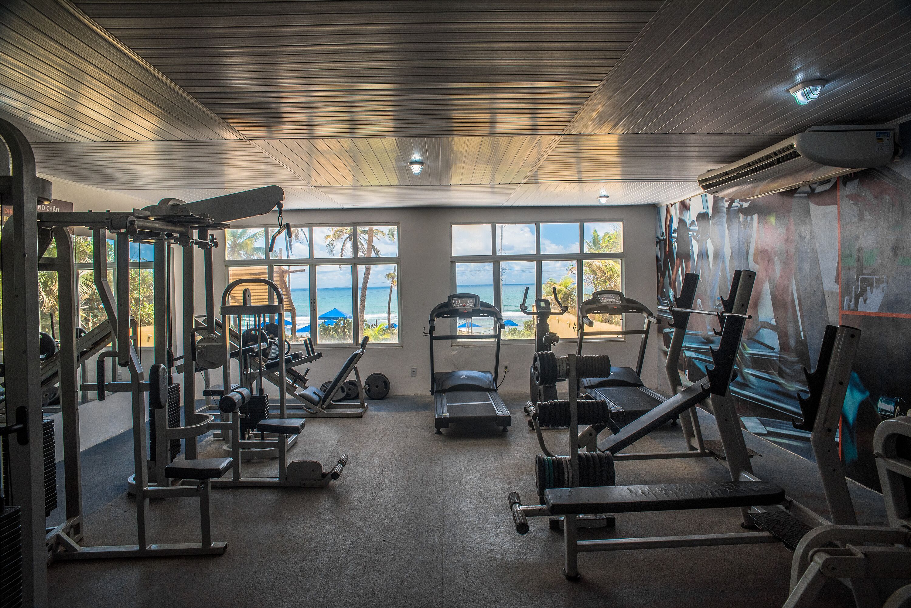 Un gimnasio con diversos equipos de ejercicio, amplias ventanas con vista a la playa y un equipo de aire acondicionado en el techo.