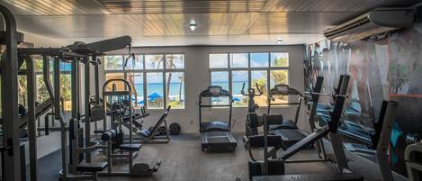Une salle de sport équipée de divers appareils d’exercice, de grandes fenêtres donnant sur la plage et d’un climatiseur installé au plafond.