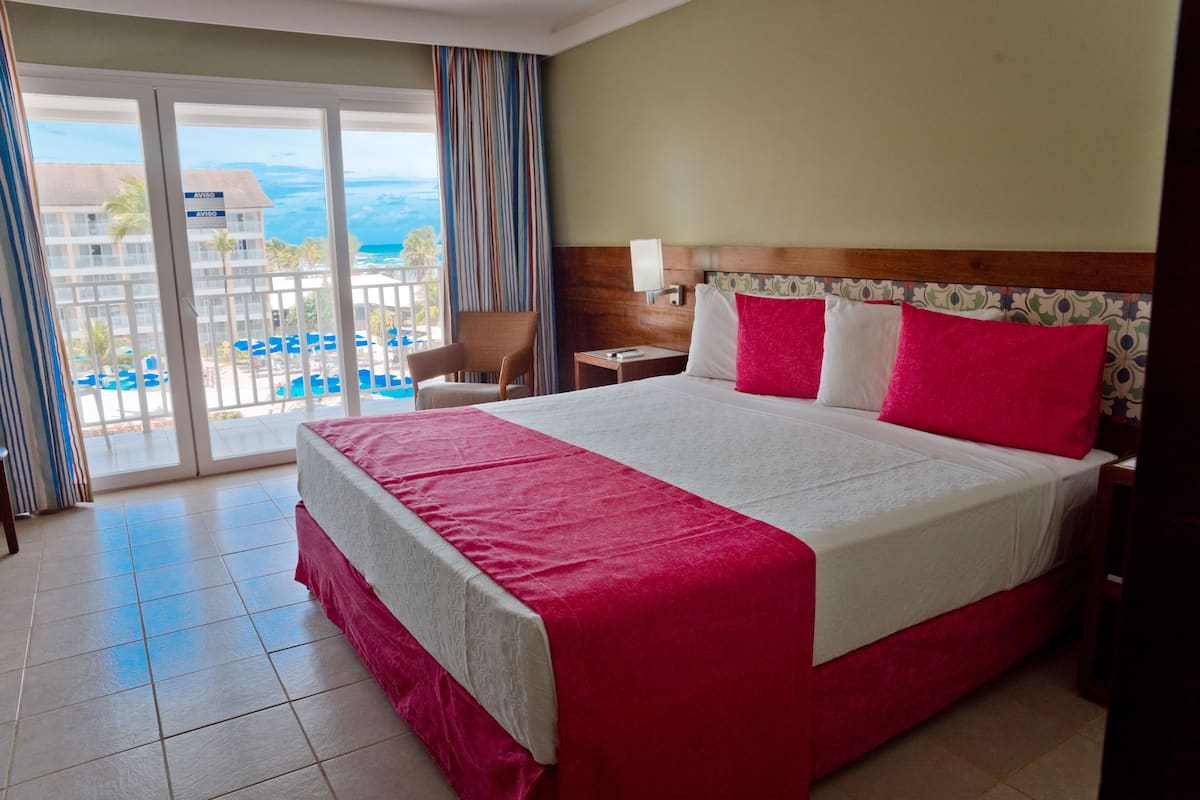 Ein Hotelzimmer mit einem großen Bett, einem Sessel, einem Nachttisch und Meerblick durch eine Schiebetür.
